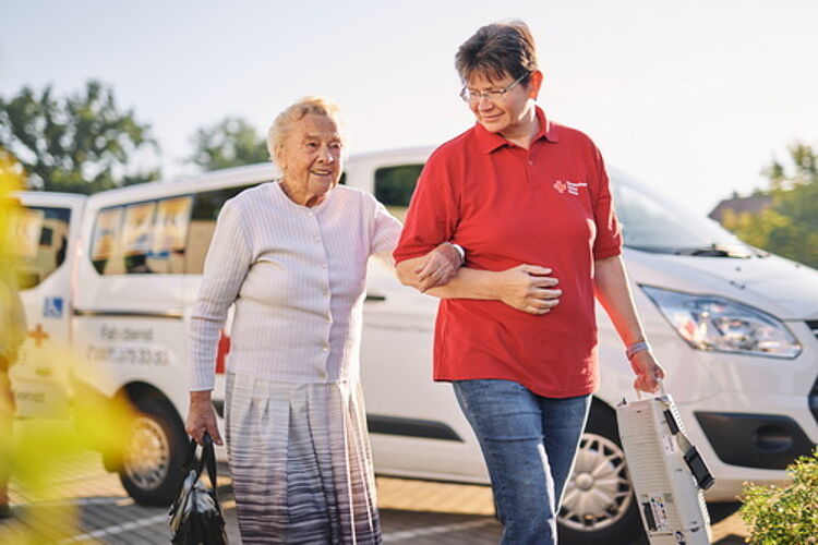 Eine DRK-Mitarbeiterin vom Fahrdienst führt eine Seniorin in den Tagestreff