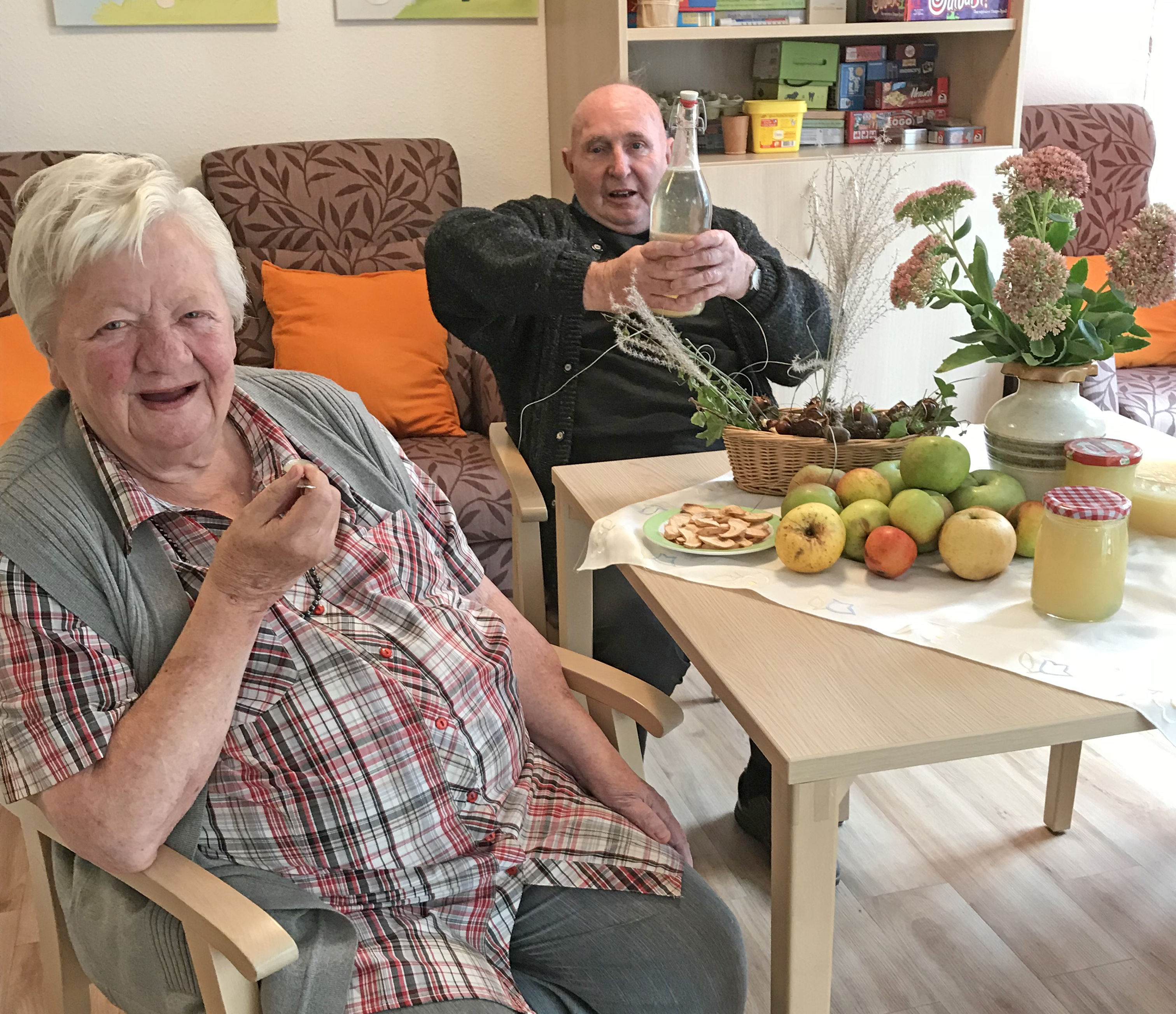 Beim Apfelfest wird jedes Jahr fleißig geschnippelt. Eine Dame und ein Herr in der Tagespflege Herzlake sitzen am Tisch und freuen sich über selbstgemachtes Apfelkompott und Apfelsaft.