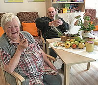Eine Dame und ein Herr in der Tagespflege Herzlake sitzen am Tisch und freuen sich über selbstgemachtes Apfelkompott und Apfelsaft.