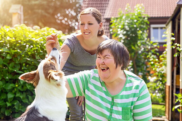 Eine Betreuungskraft und eine unterstützungsbedürftige Frau spielen mit einem Hund